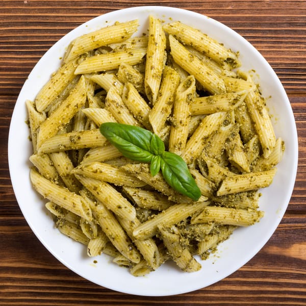 Penne pasta with vibrant green pesto sauce and a fresh basil garnish in a white bowl.