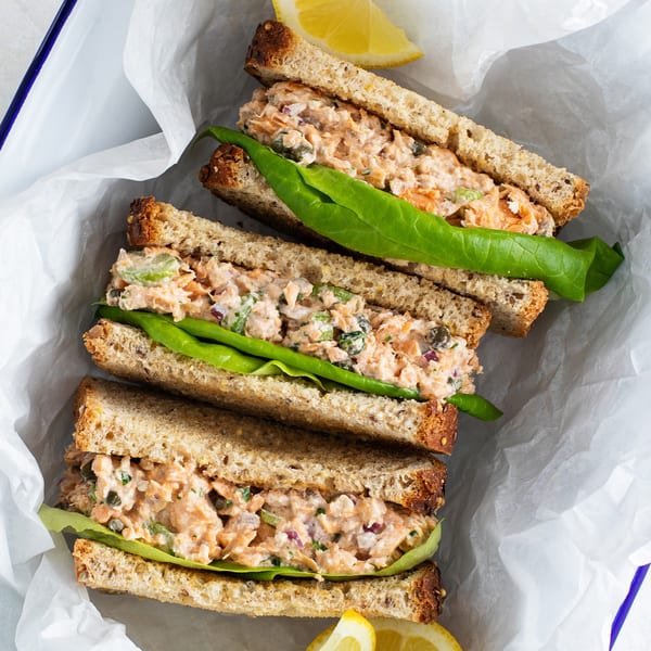 Three salmon salad sandwiches on whole wheat bread with lettuce and lemon wedges in a white container.